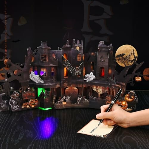 Hand writing on paper next to a spooky haunted house decoration with glowing lights and Halloween motifs.